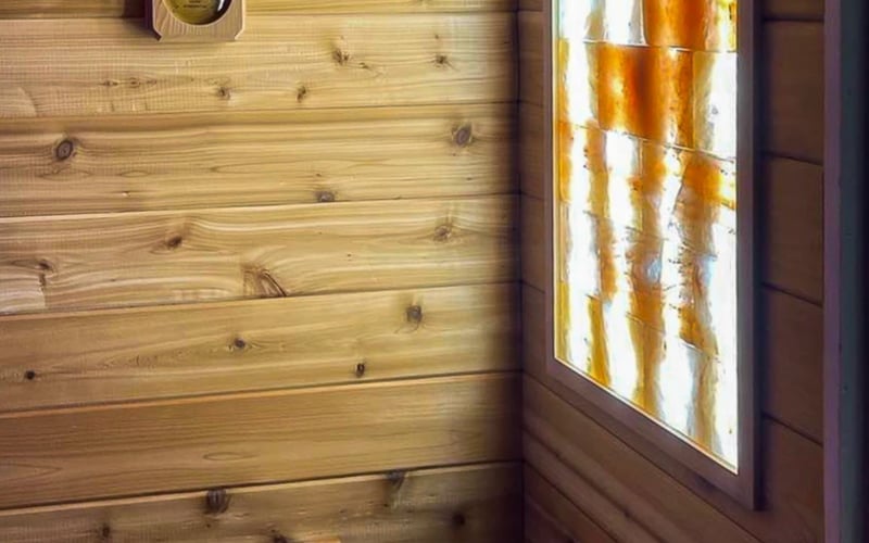 Sauna interior with light wood, heater filled with rocks, brass faucet, and stained glass window